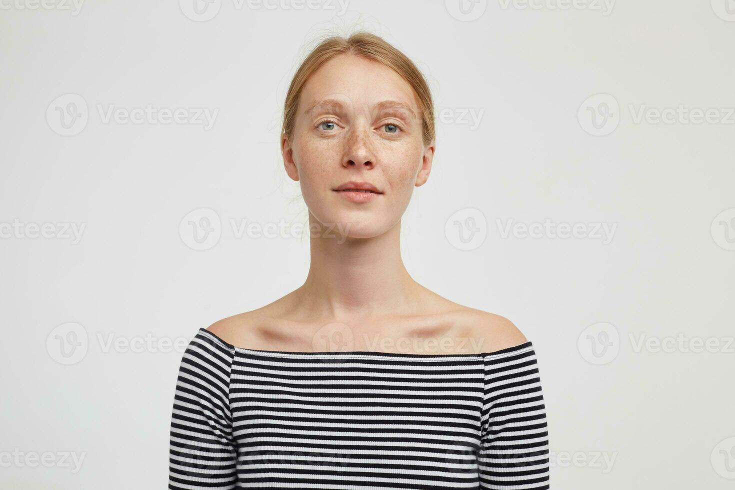 Studio photo of beautiful young redhead female with casual hairstyle looking into camera and keeping her hands down while standing over white background