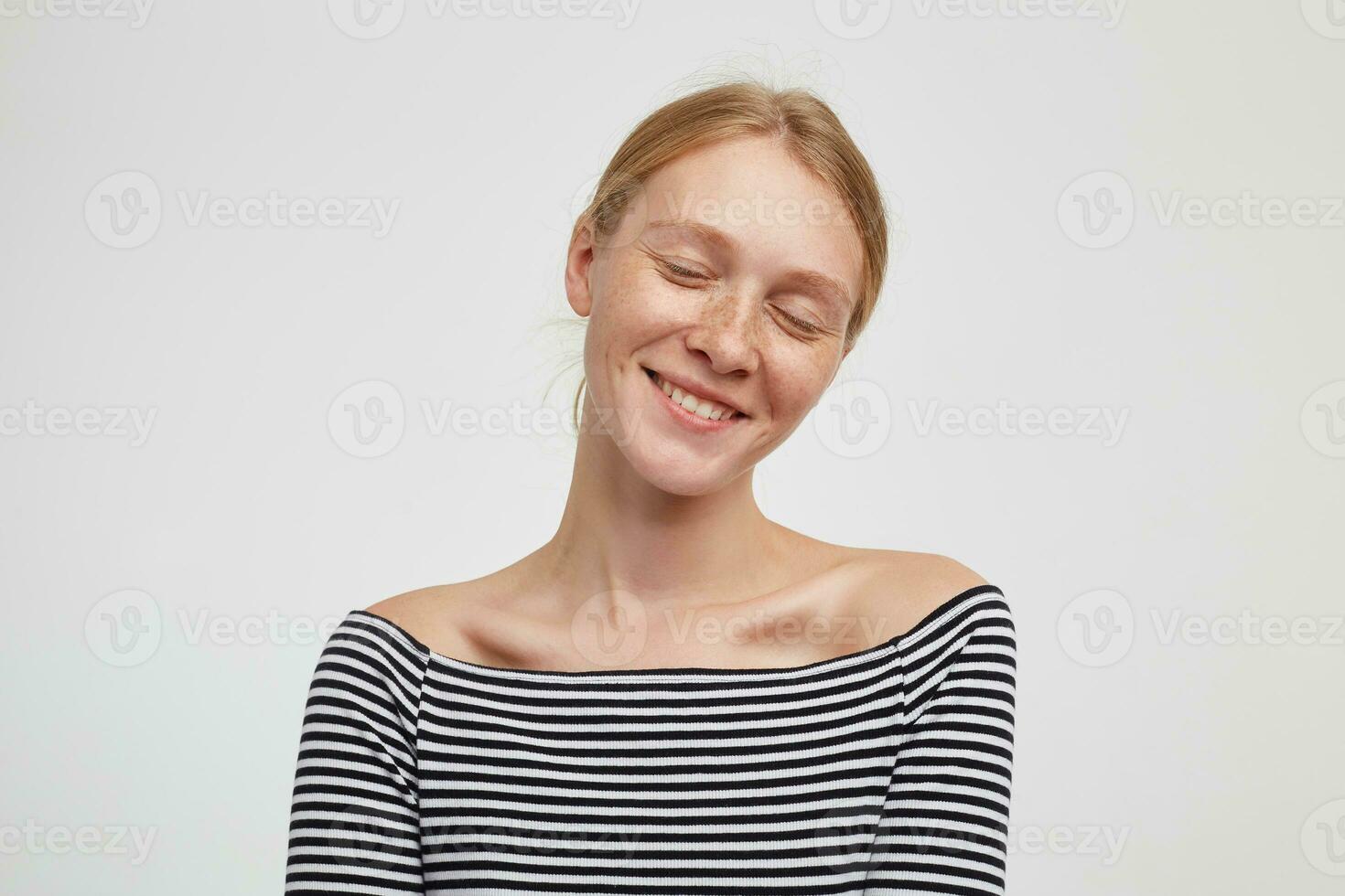 Pleasant looking young cheerful redhead female with casual hairstyle smiling nicely with closed eyes while posing over white background in casual wear photo