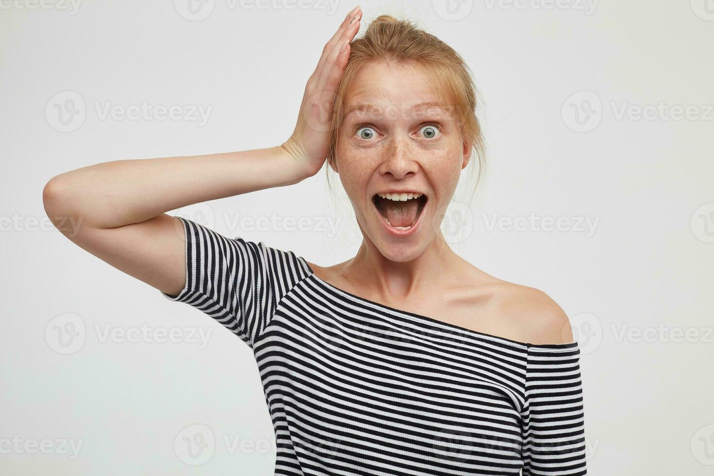 Indoor shot of overjoyed young lovely redhead female dressed in casual wear holding raised hand on her head and looking excitedly at camera with wide mouth opened, isolated over white background photo