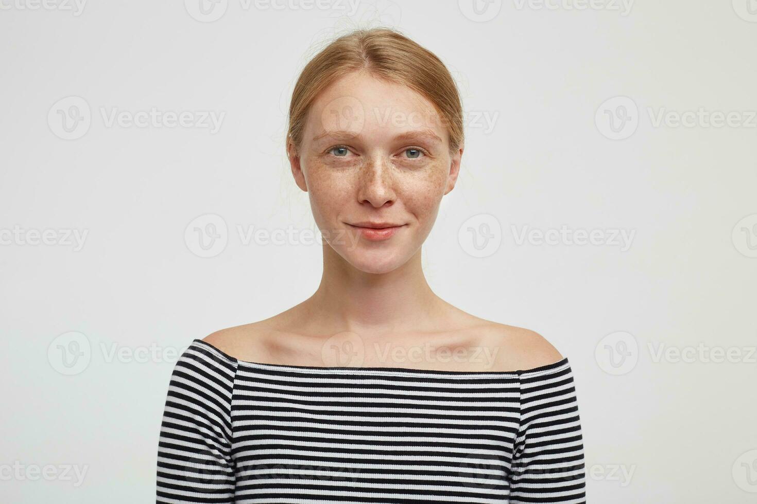 Portrait of positive young green-eyed lovely redhead woman looking at camera with pleasant smile while standing over white background with hands down photo