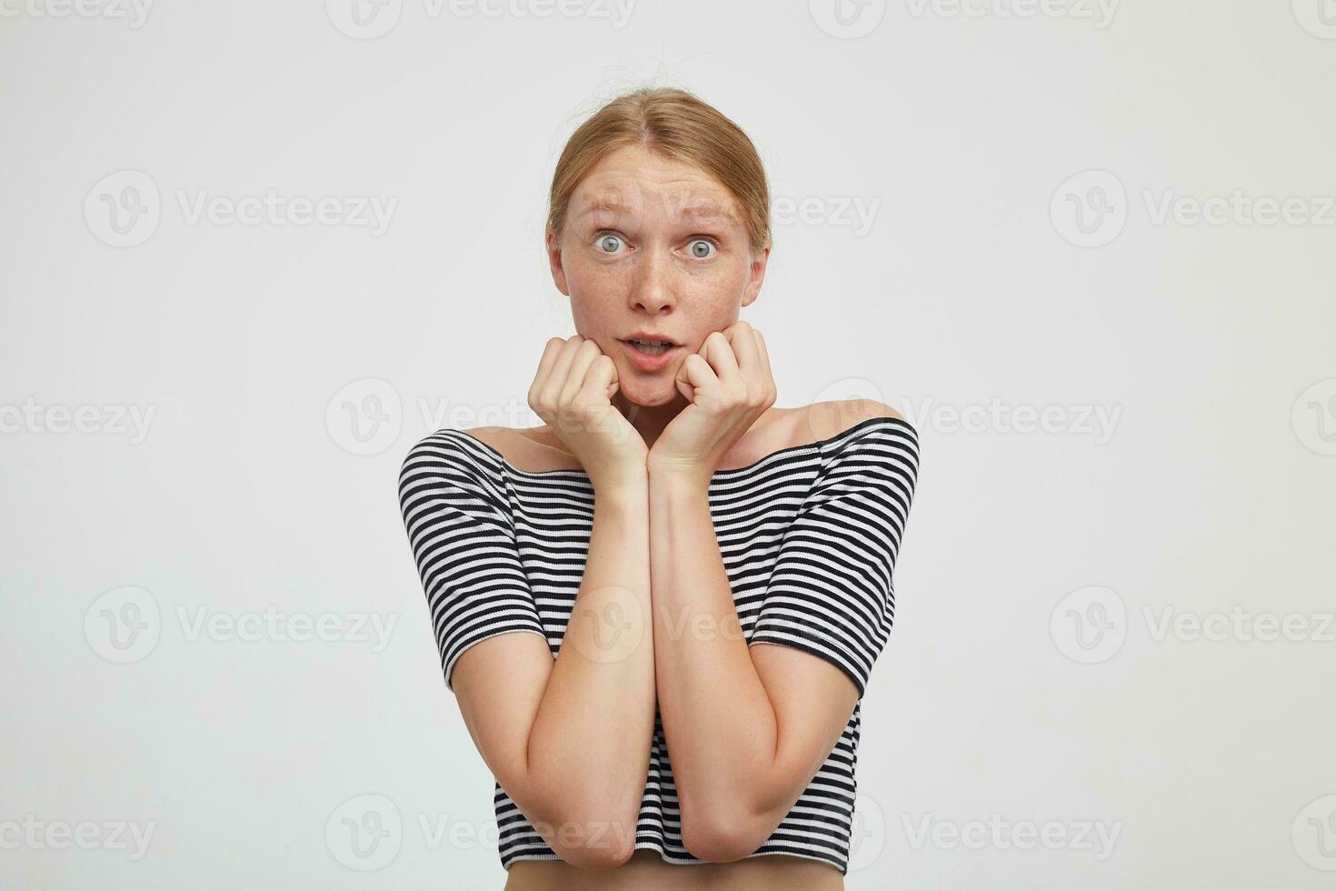 Scared young green-eyed attractive woman with casual hairstyle looking frightened at camera and keeping raised hands on her cheeks, isolated over white background photo