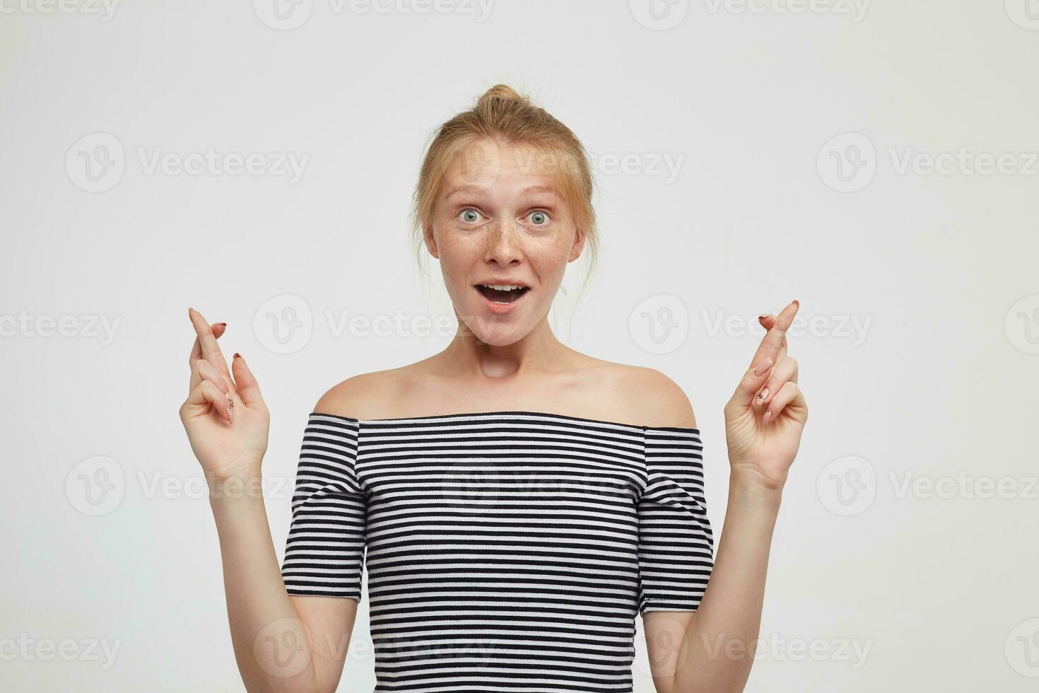 Agitated young attractive redhead female with bun hairstyle keeping her fingers crossed while looking excitedly at camera with wide eyes and mouth opened, posing over white background photo