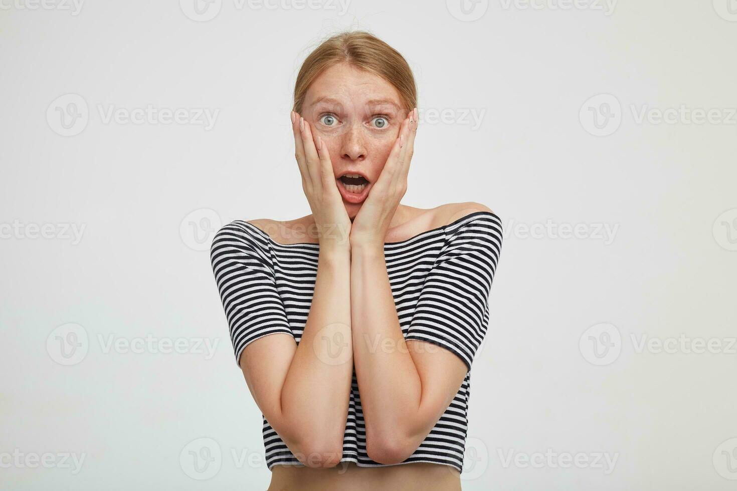 Afraid young pretty redhead female dressed in casual wear holding her face with raised palms and looking at camera with opened mouth, posing over white background photo