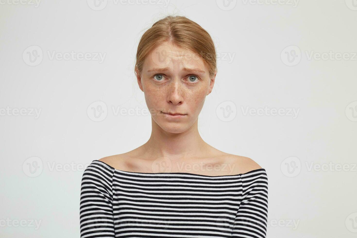 Unhappy young attractive redhead woman with casual hairstyle biting her lips while looking sadly at camera, keeping hands down while posing over white background photo