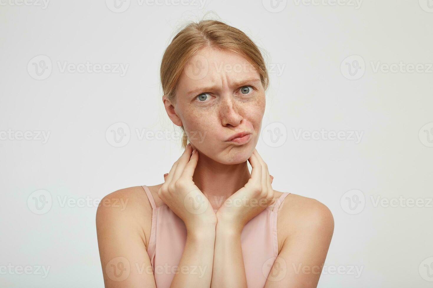Puzzled young attractive redhead lady frowning her eyebrows and twisting mouth while looking confusedly at camera, keeping raised hands on neck while posing over white background photo
