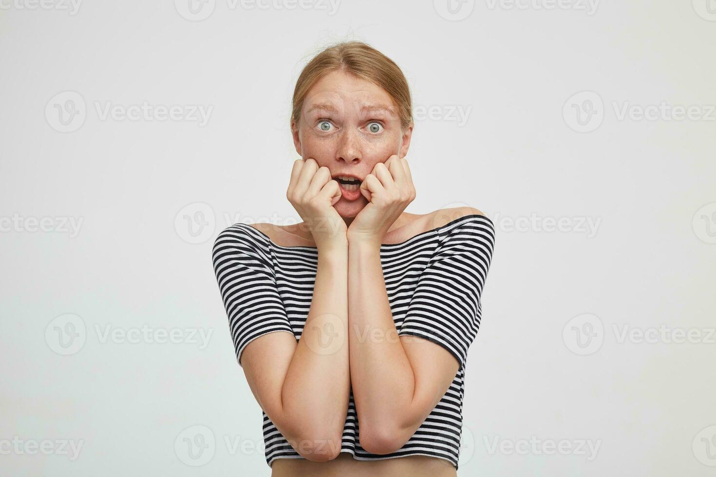 Frightened young lovely redhead lady holding raised hands on her face and looking scaredly at camera with wide eyes opened, isolated over white background photo