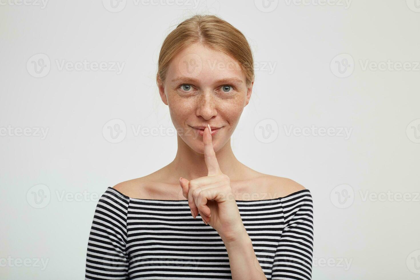 Portrait of young lovely redhead female holding index finger on her lips while asking to keep silence and smiling cunningly, standing against white background photo