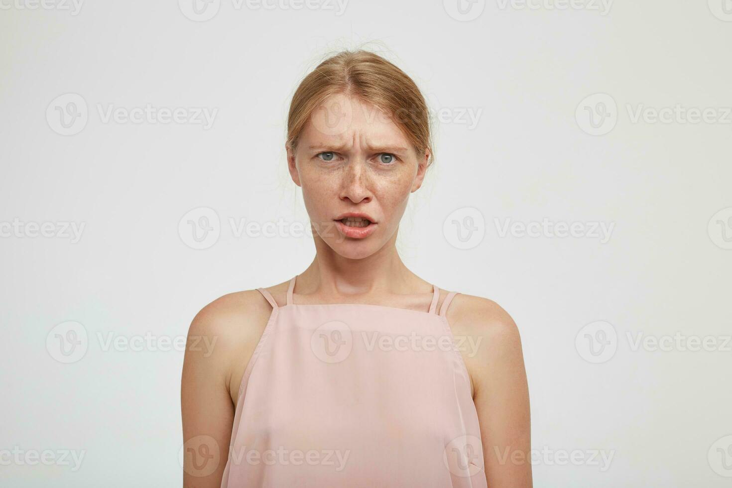 Confused young green-eyed redhead female grimacing her face while looking perplexedly at camera and frowning her eyebrows, isolated over white background photo