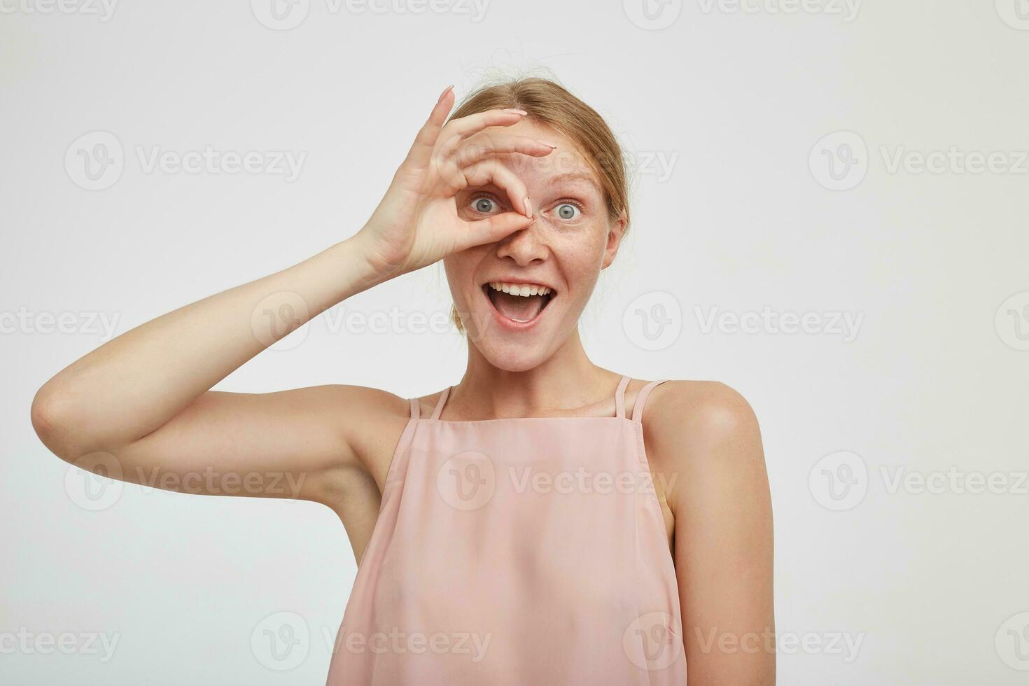 Funny shot of young attractive redhead female with casual hairstyle raising ok gesture to her eye and looking joyfully at camera with opened mouth, isolated over white background photo