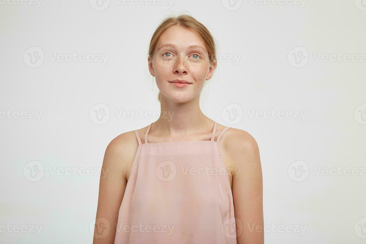 Portrait of young green-eyed pretty redhead woman looking calmly in front of herself and smiling gently, dressed in pink top while standing over white background photo