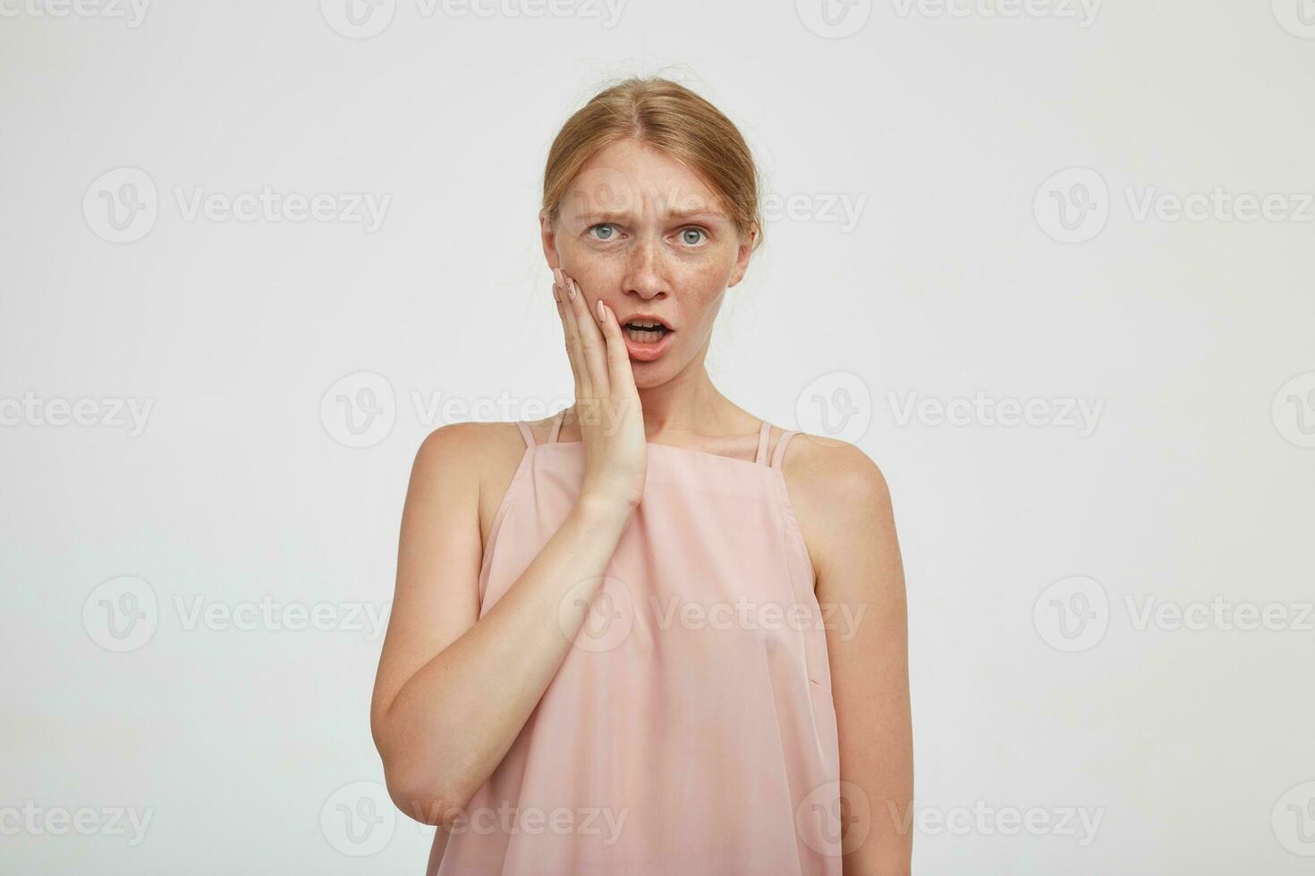 Bewildered young pretty redhead woman with casual hairstyle holding palm on her face while looking confusedly at camera and frowning her eyebrows, standing over white background photo