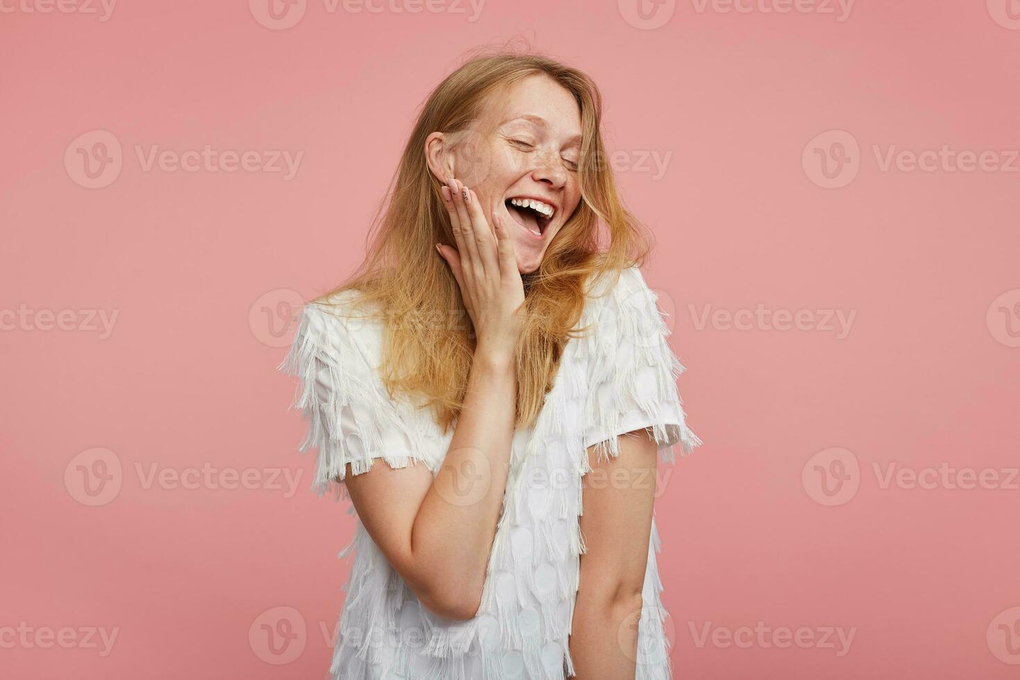 Indoor shot of joyful attractive young redhead lady with wild hairstyle holding palm on her cheek while laughing happily with closed eyes, standing over pink background photo