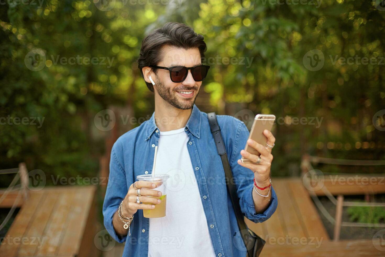 Young pretty bearded male with dark hair walking through green city garden, drinking lemonade and holding mobile phone in hand, looking at screen and smiling cheerfully photo
