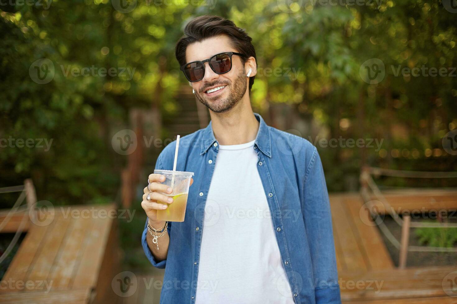 Happy attractive dark haired man in blue shirt standing over green trees on sunny day, having good day and drinking lemonade in outdoor public place photo