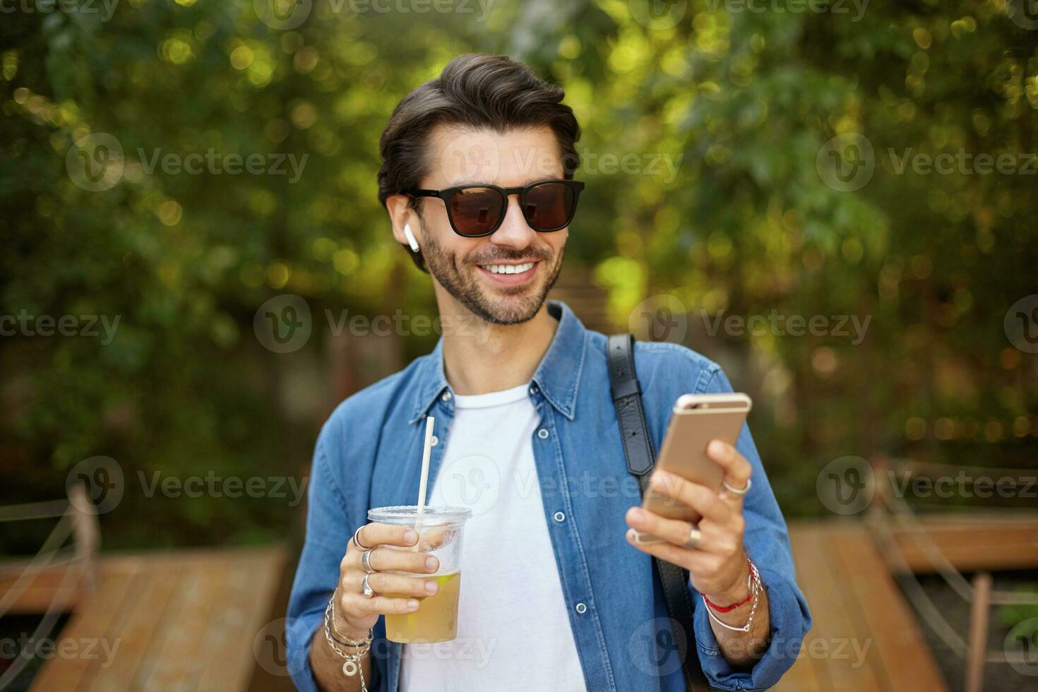 Outdoor portrait of handsome dark haired male with smartphone in hand, reading good news and having nice mood, drinking ice tea in plastic cup photo