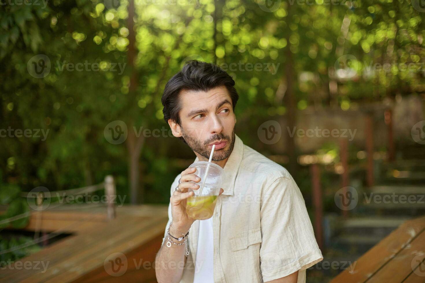 Outdoor shot of young pretty male with beard sitting over green trees on bench, looking aside and drinking lemonade, wearing casual clothes photo