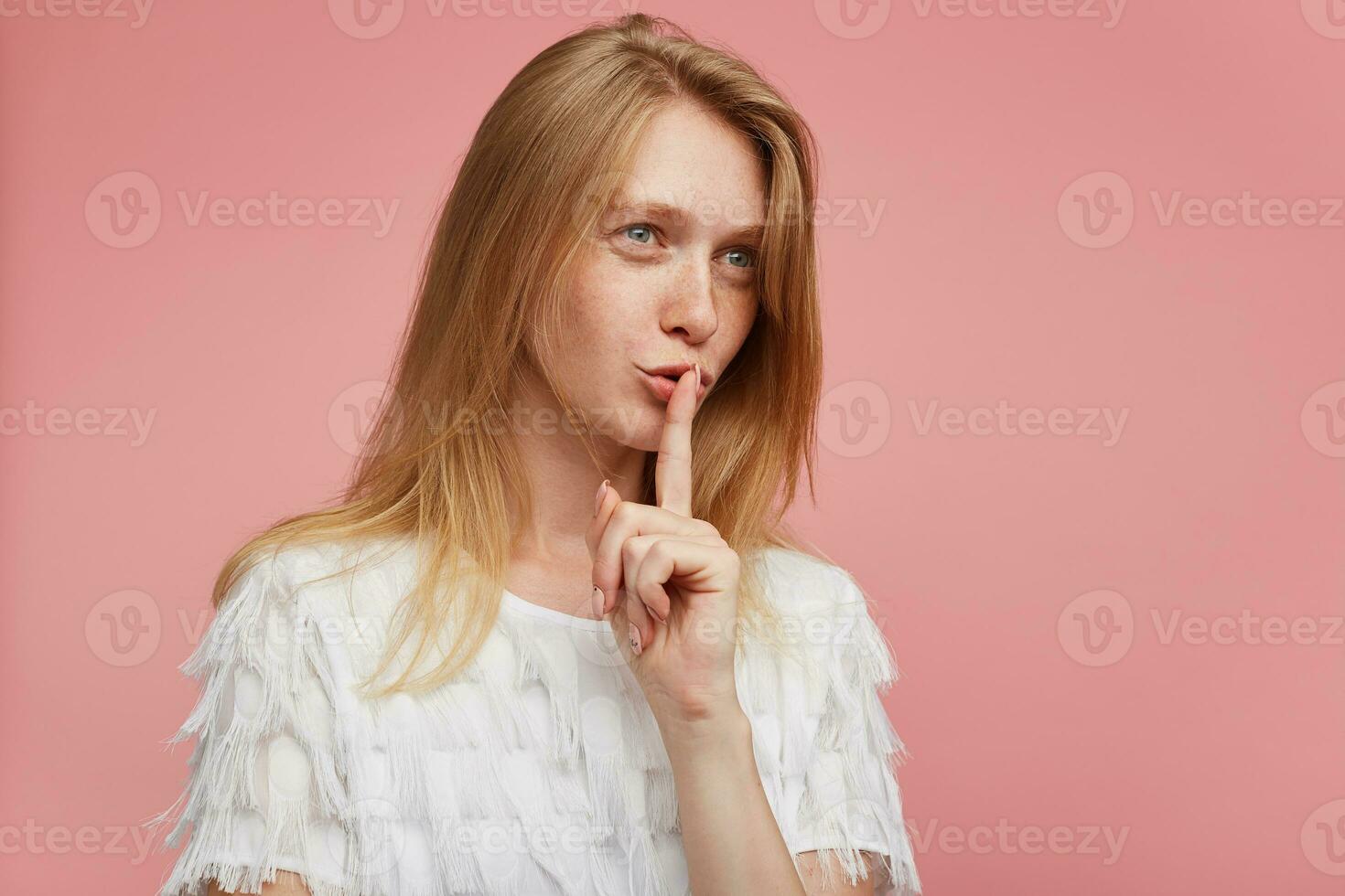 Side view of lovely young redhead lady with casual hairstyle raising hand with hush gesture and asking to keep secret, standing against pink background in white shirt photo