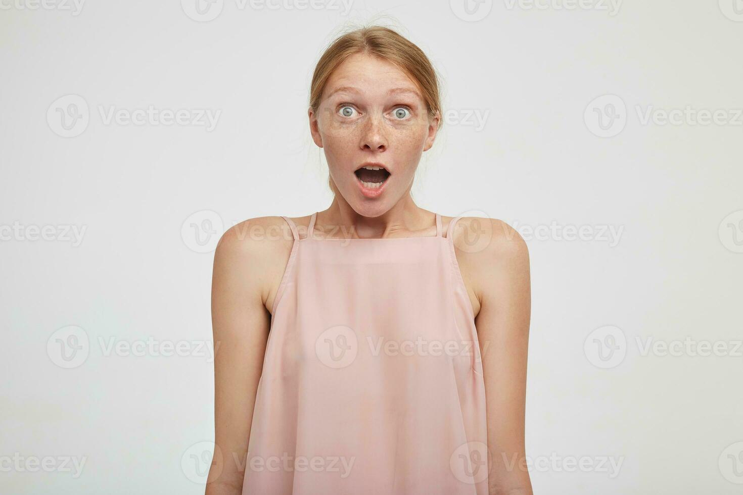 Astonieshed young pretty redhead female rounding her green eyes while looking amazedly at camera with wide mouth opened, isolated over white background photo