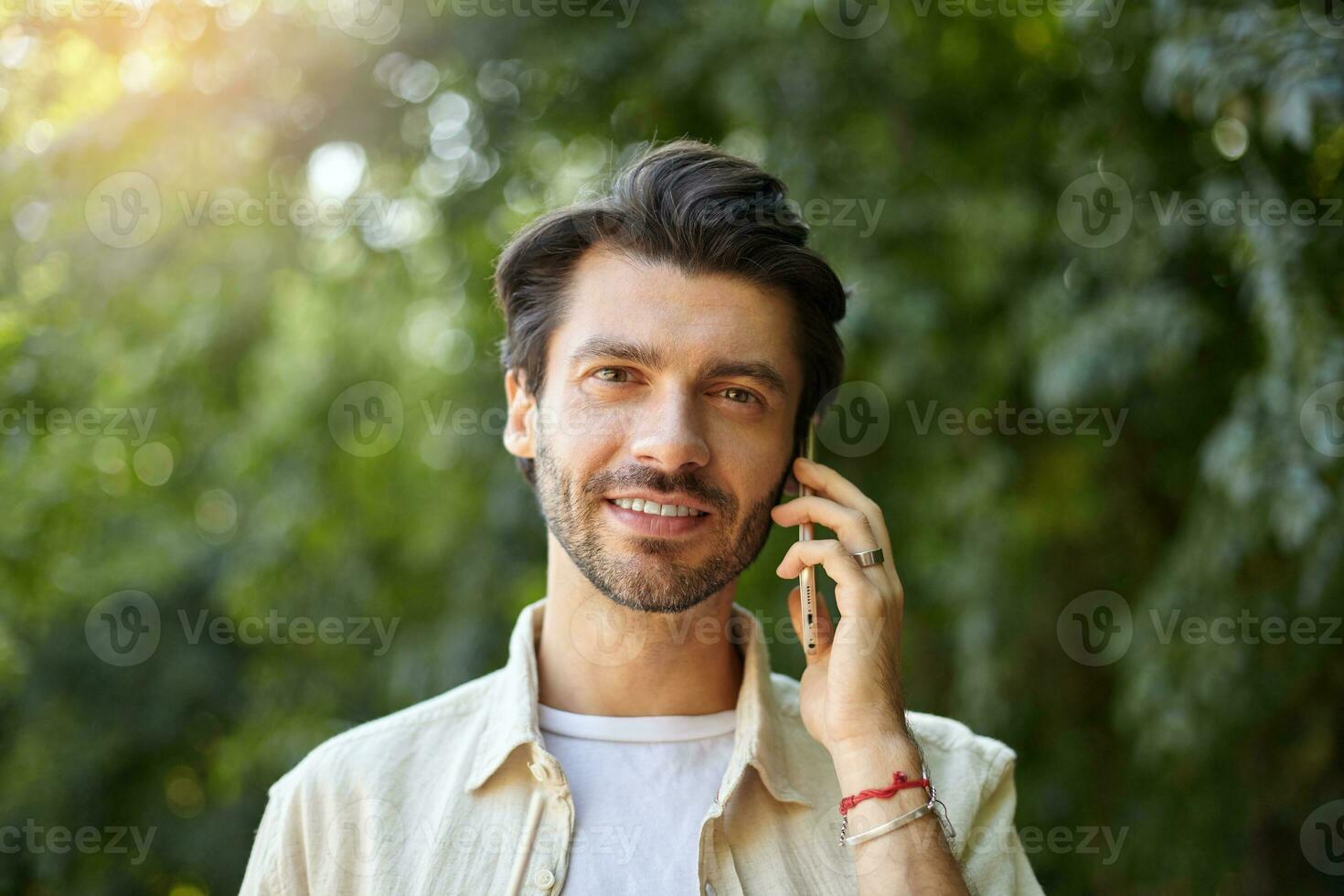 Outdoor close-up of charming young dark haired man with beard having talk on his mobile phone while walking along green garden on sunny day photo