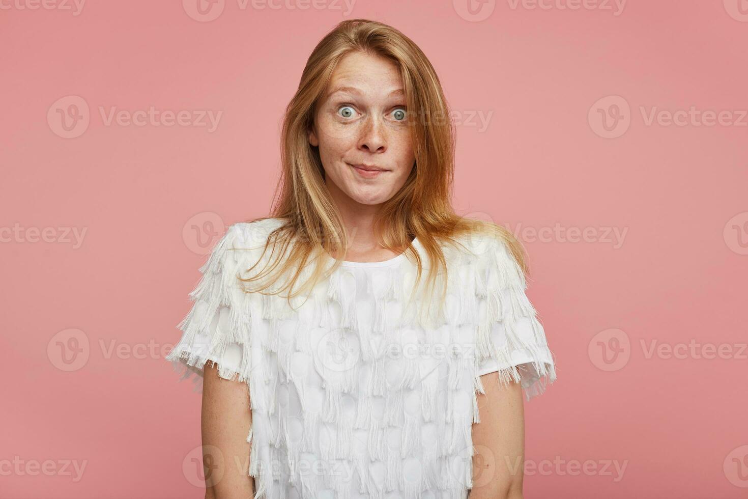 Amazed young pretty redhead woman with casual hairstyle rounding her green-gray eyes while looking surprisedly to camera, isolated over pink background with hands down photo