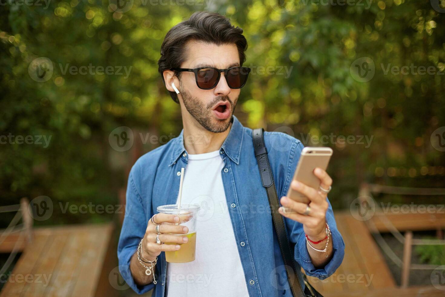 Surprised attractive young bearded man in sunglasses standing over green park with mobile phone in hand, receiving unexpected news, wearing casual clothes photo