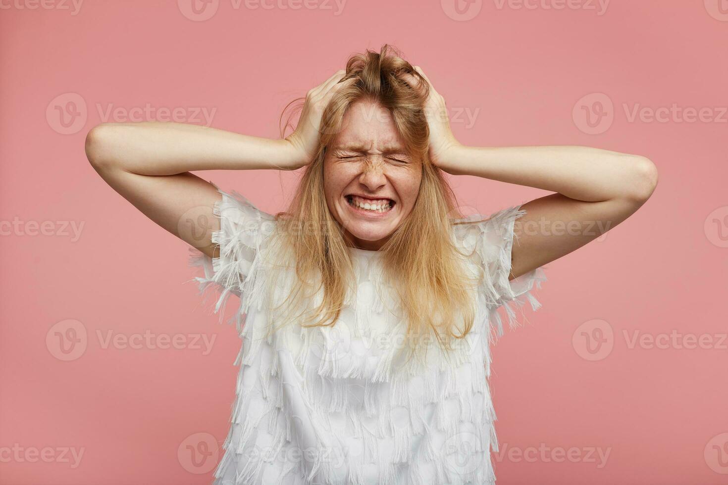 Stressed young redhead lady frowning her face and keeping eyes closed while clutching her head with raised hands, wearing white festive blouse while posing over pink background photo