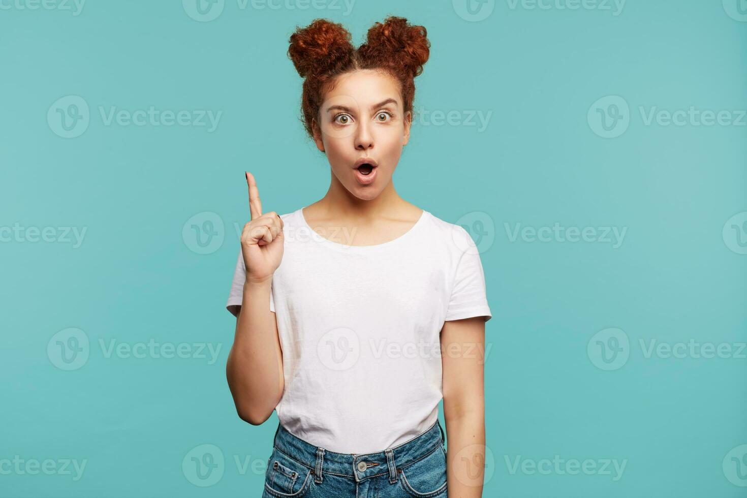 Open-eyed young brown haired curly woman raising hand as she is having idea and looking at camera with agitated face, dressed in white basic t-shirt and jeans while posing over blue background photo