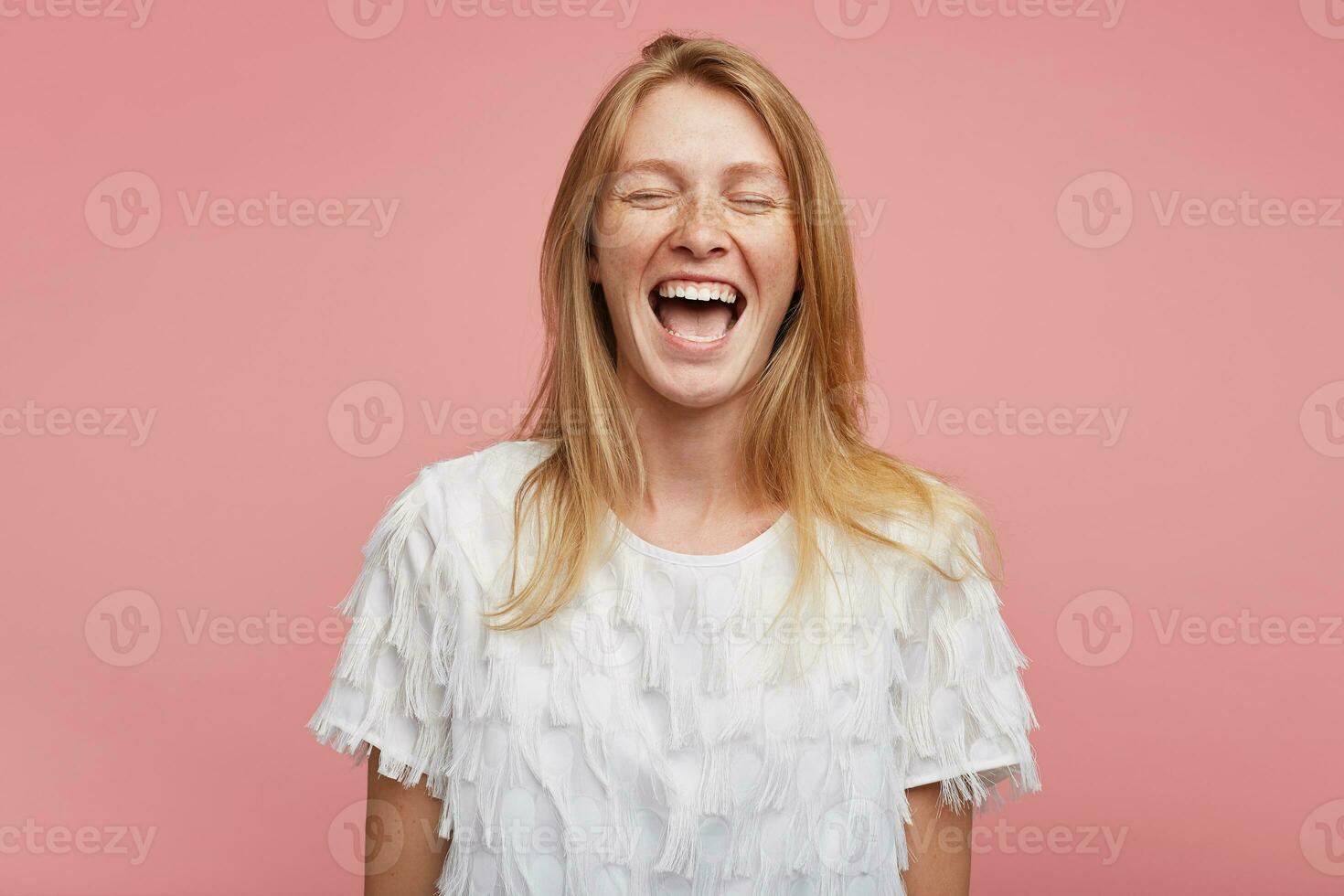Good looking overjoyed young attractive female with foxy hair keeping her mouth wide opened while laughing happily with closed eyes, standing against pink background photo