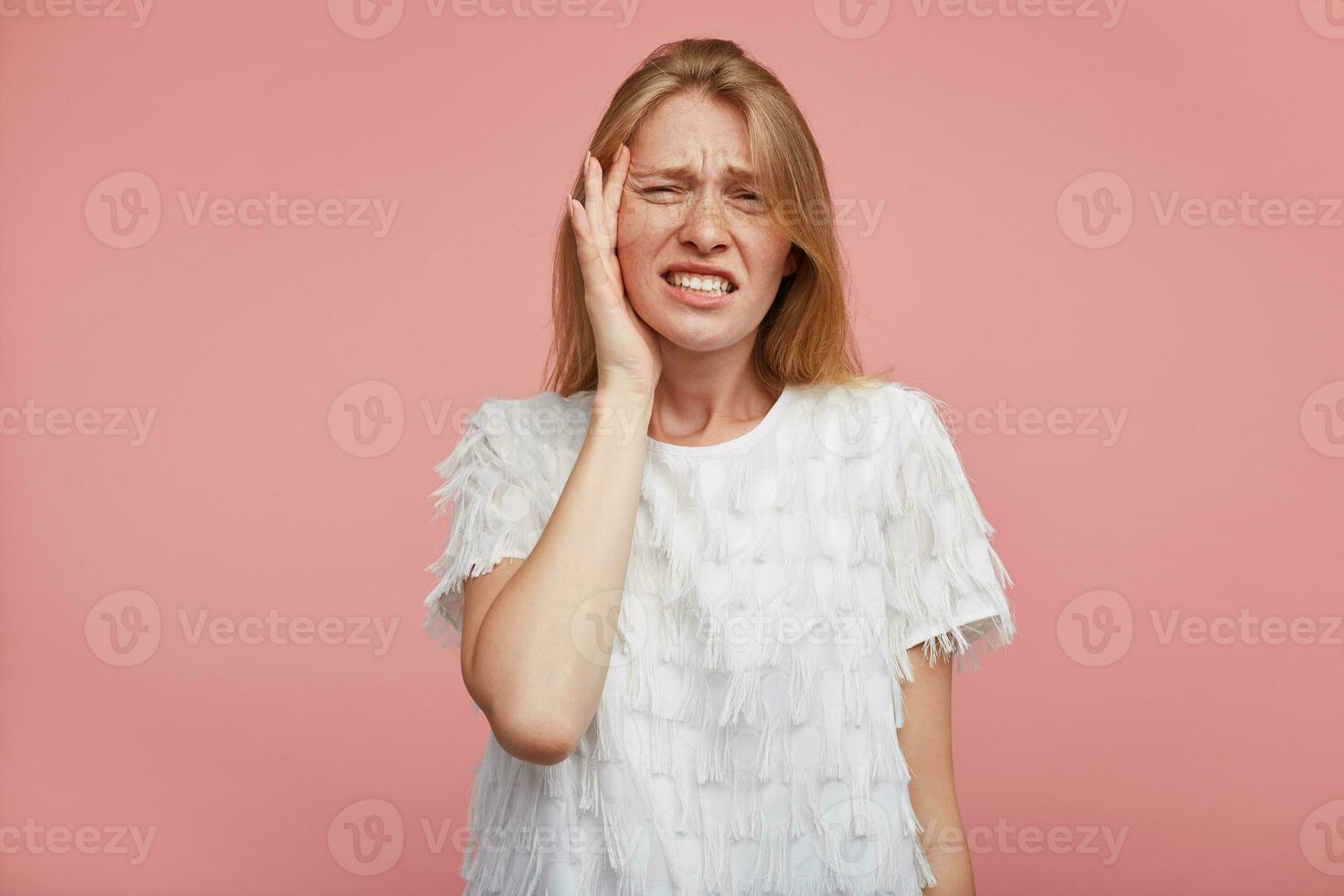 Portrait of sad young pretty woman with foxy hair showing her teeth while looking unhappily at camera and holding raised hand on her face, standing over pink background photo