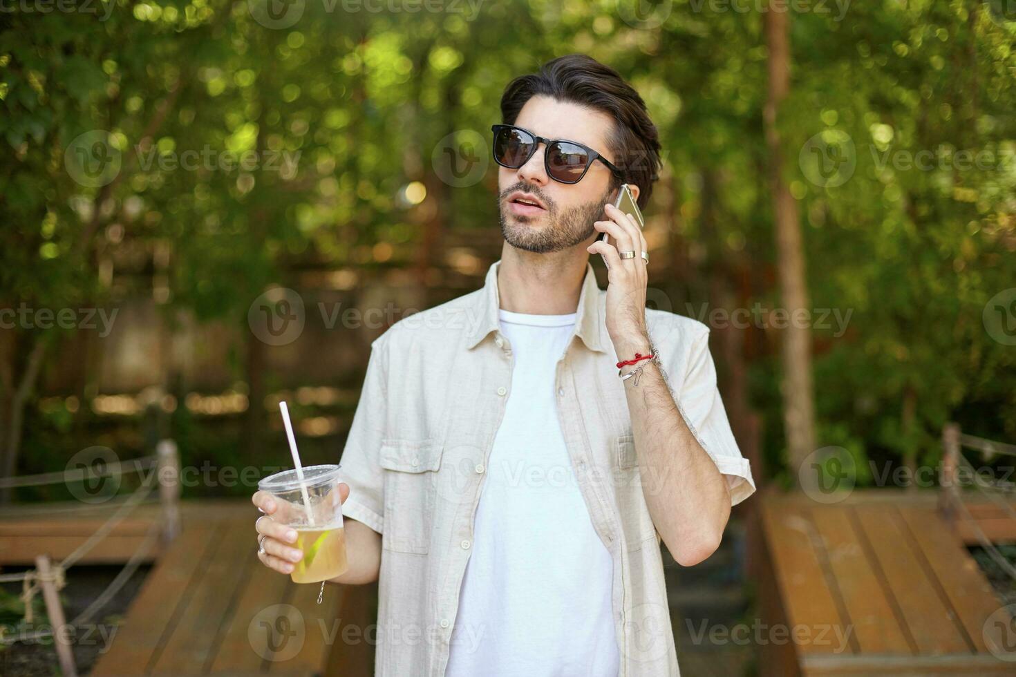 Young attractive bearded man in beige shirt and sunglasses making call with his mobile phone, walking around green city prk with lemonade in hand photo