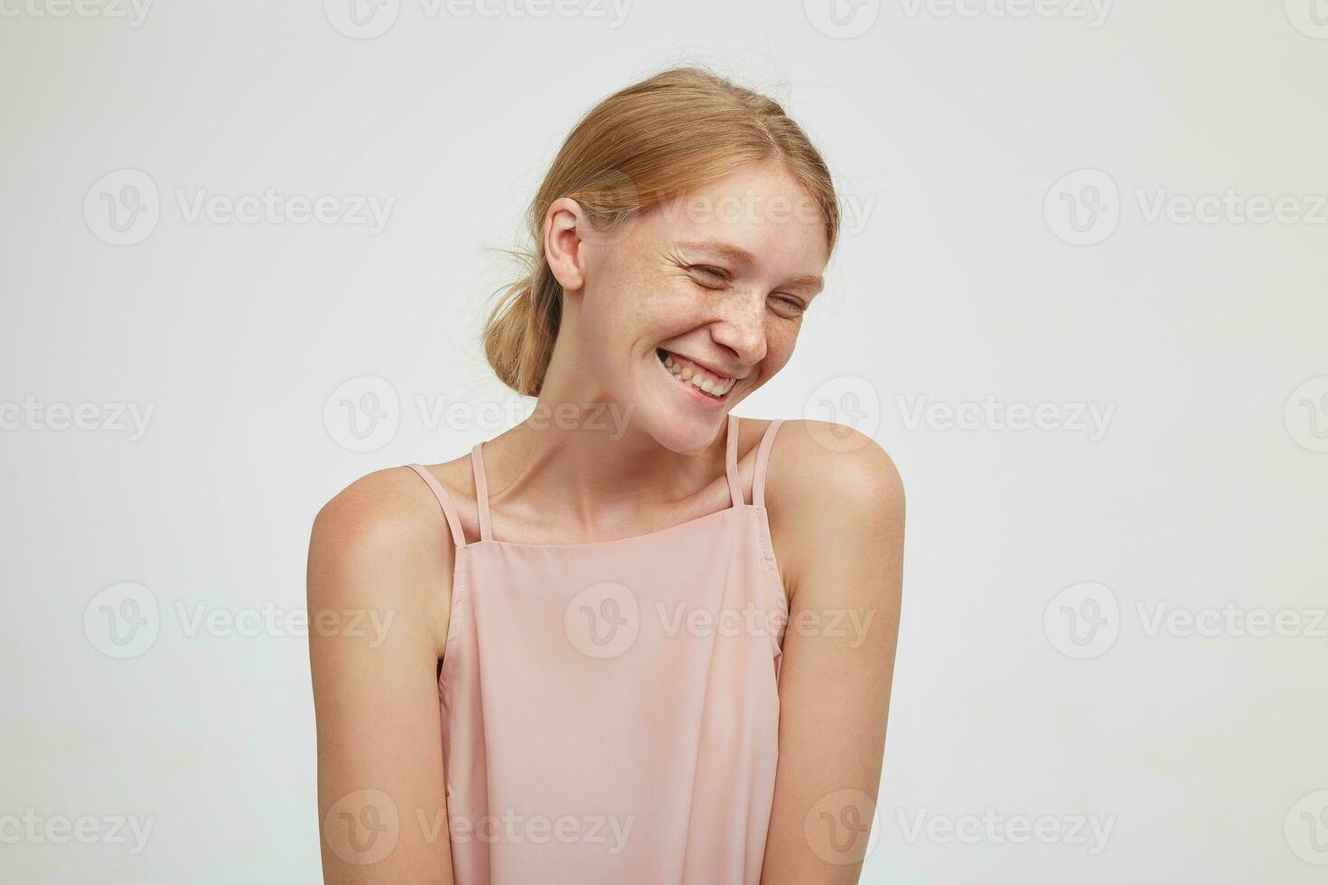 Pleased young beautiful redhead female dressed in pink shirt being in nice mood and smiling cheerfully, keeping her hands down while posing over white background photo