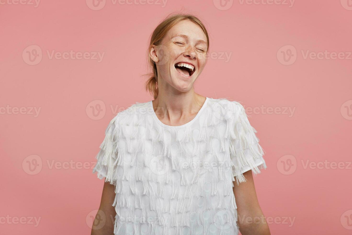 Pleasant looking young happy redhead woman with ponytail hairstyle keeping her eyes closed while laughing joyfully, being in high spirit while standing over pink background photo