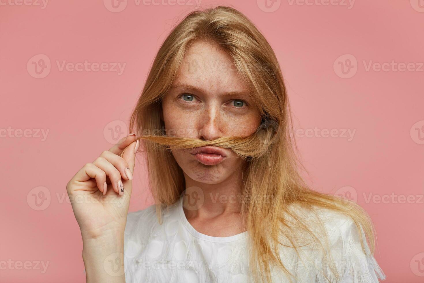 Closeup of young pretty redhead female with casual hairstyle imitating mustache with lock of hair and looking seriously at camera, dressed in casual clothes while posing over pink background photo