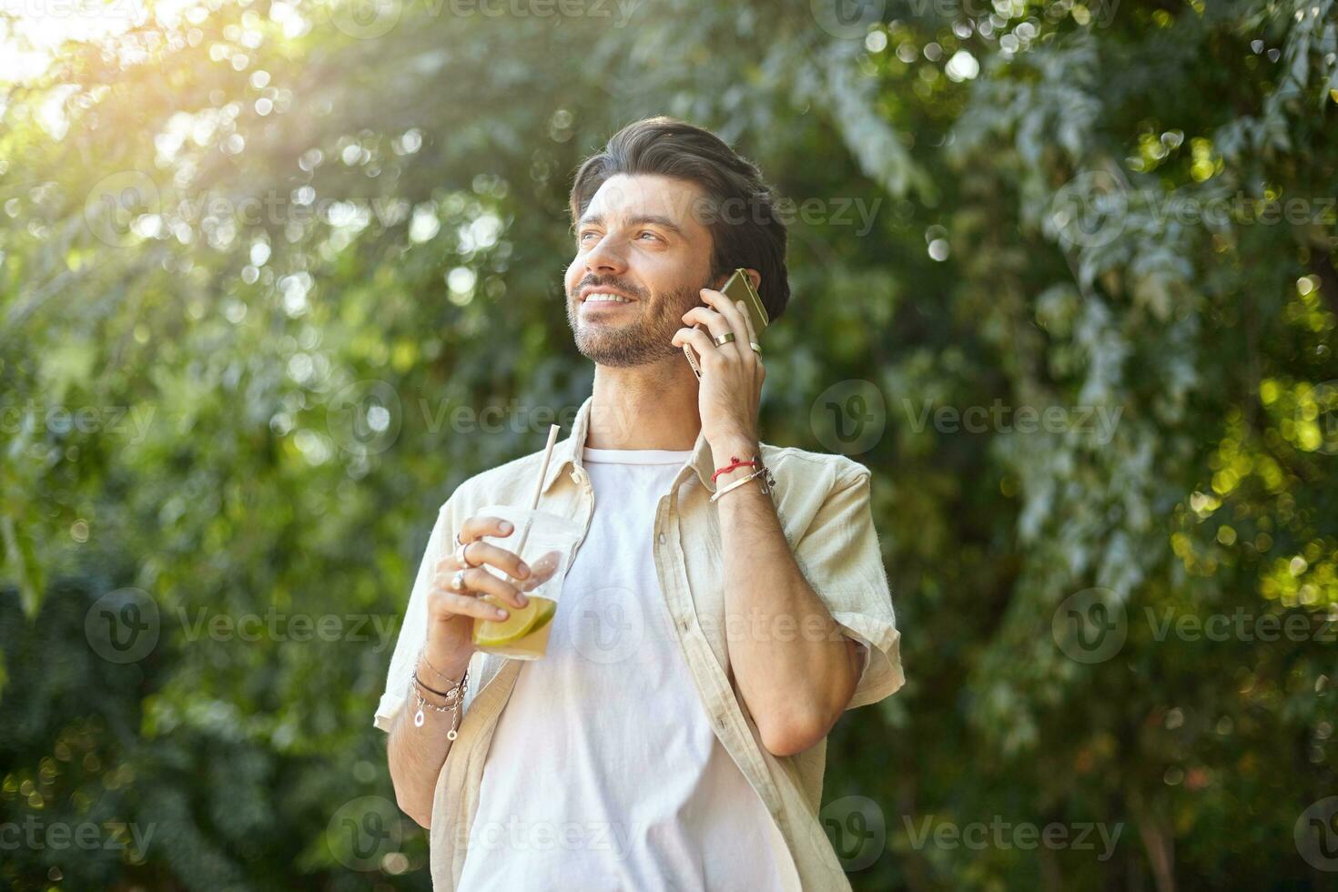 positivo joven bonito hombre con barba posando terminado verde ciudad parque en calentar soleado día, haciendo llamada con su teléfono inteligente y participación el plastico taza con limonada foto