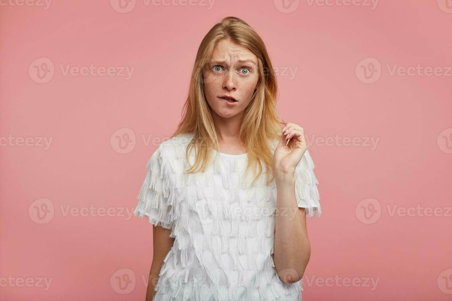 Confused young pretty female with foxy hair frowning her eyebrows and biting underlip while standing over pink background with raised hand, dressed in elegant clothes photo
