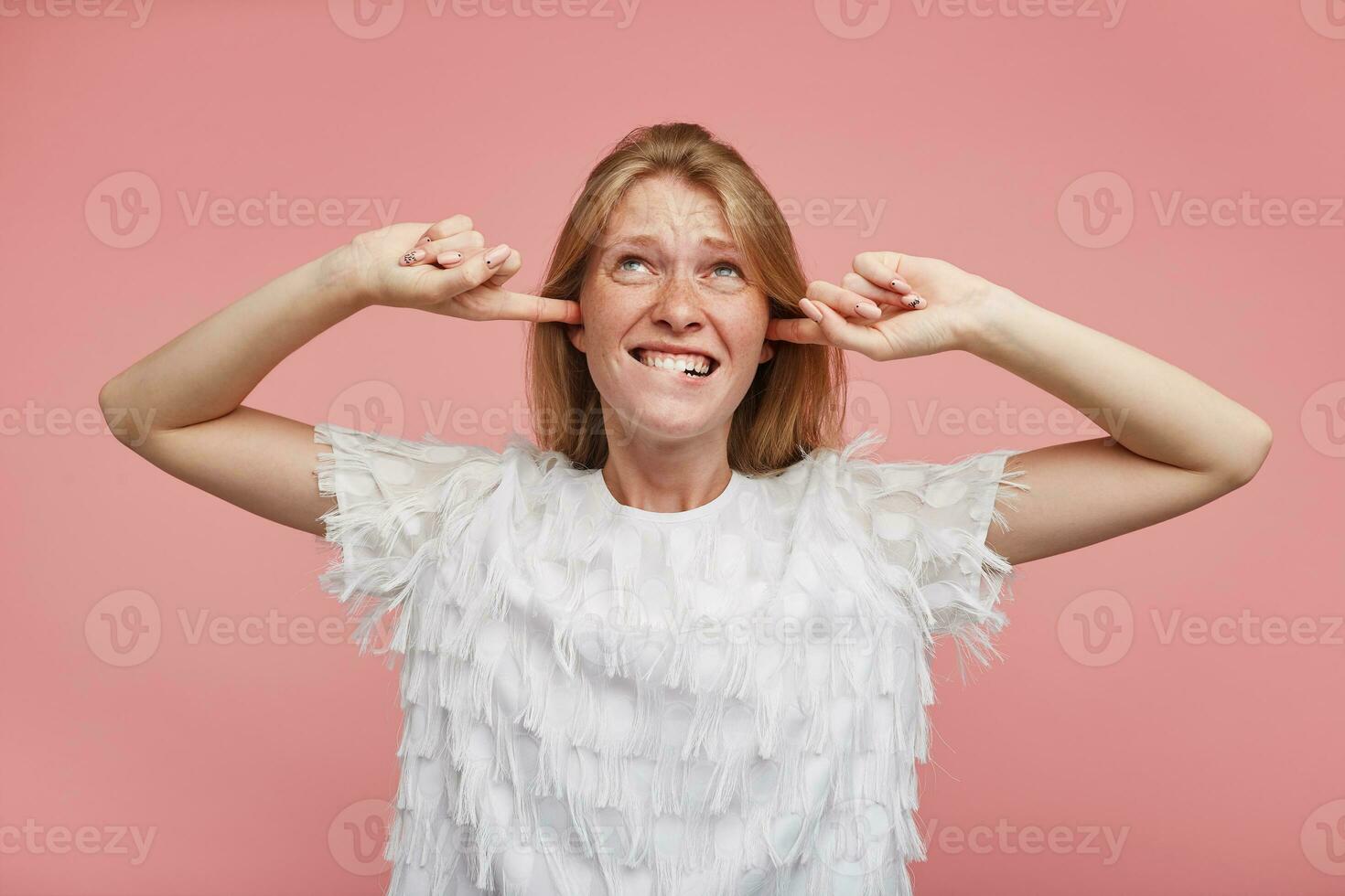 Displeased young attractive redhead lady with casual hairstyle keeping forefingers in her ears while trying to avoid annoying sounds, biting underlip while standing over pink background photo