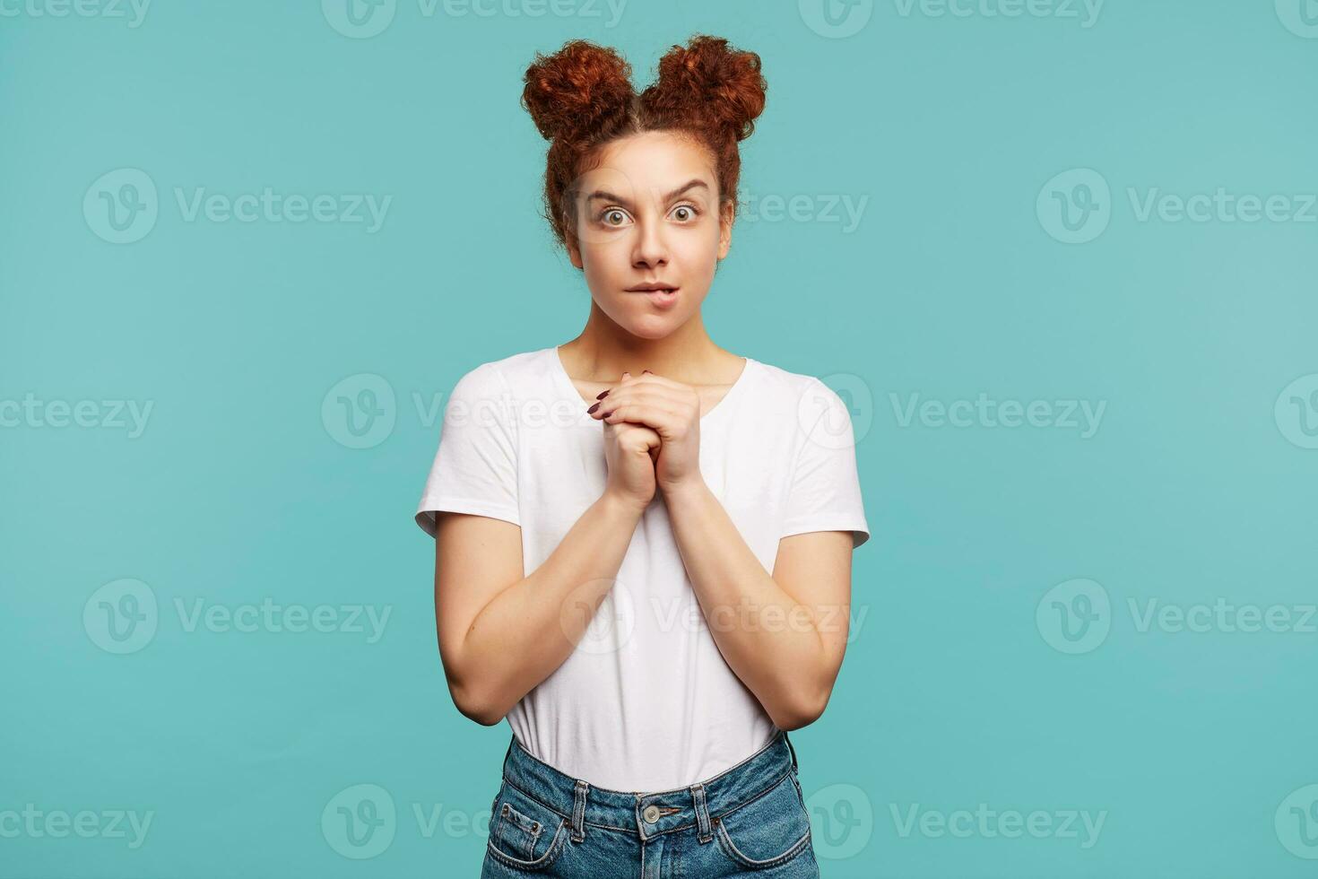 Confused young pretty curly brunette female with natural makeup keeping raised hands together and biting underlip while looking worringly at camera, standing over blue background photo