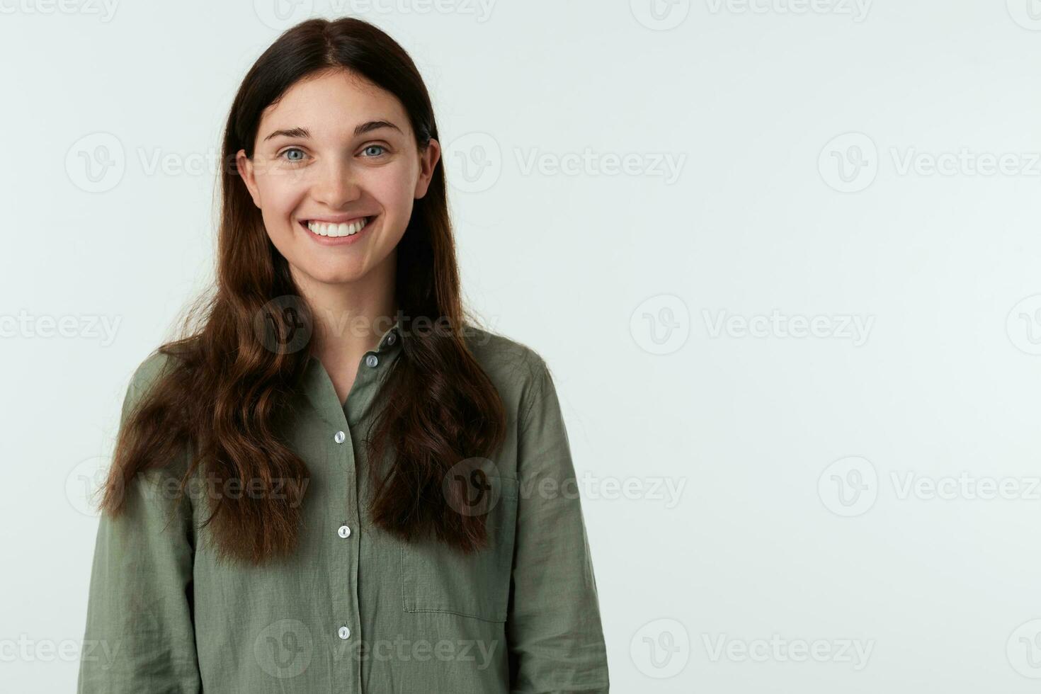 Indoor shot of young joyful brown haired lady with casual hairstyle smiling widely while looking gladly at camera, standing over white background in green shirt photo