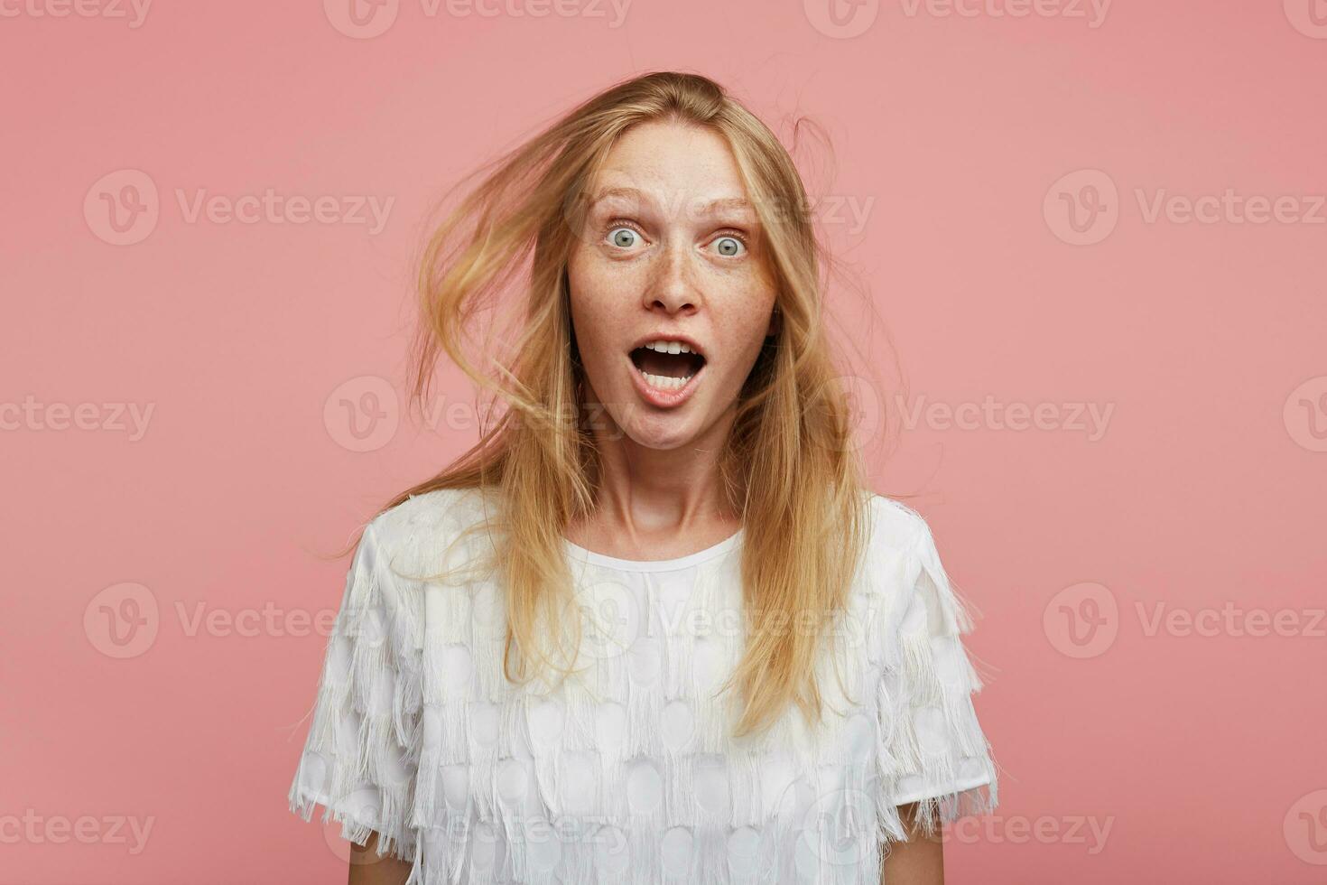 Agitated young pretty redhead female with wild hair looking excitedly at camera with wide eyes opened, raising surprisedly eyebrows while posing over pink background photo