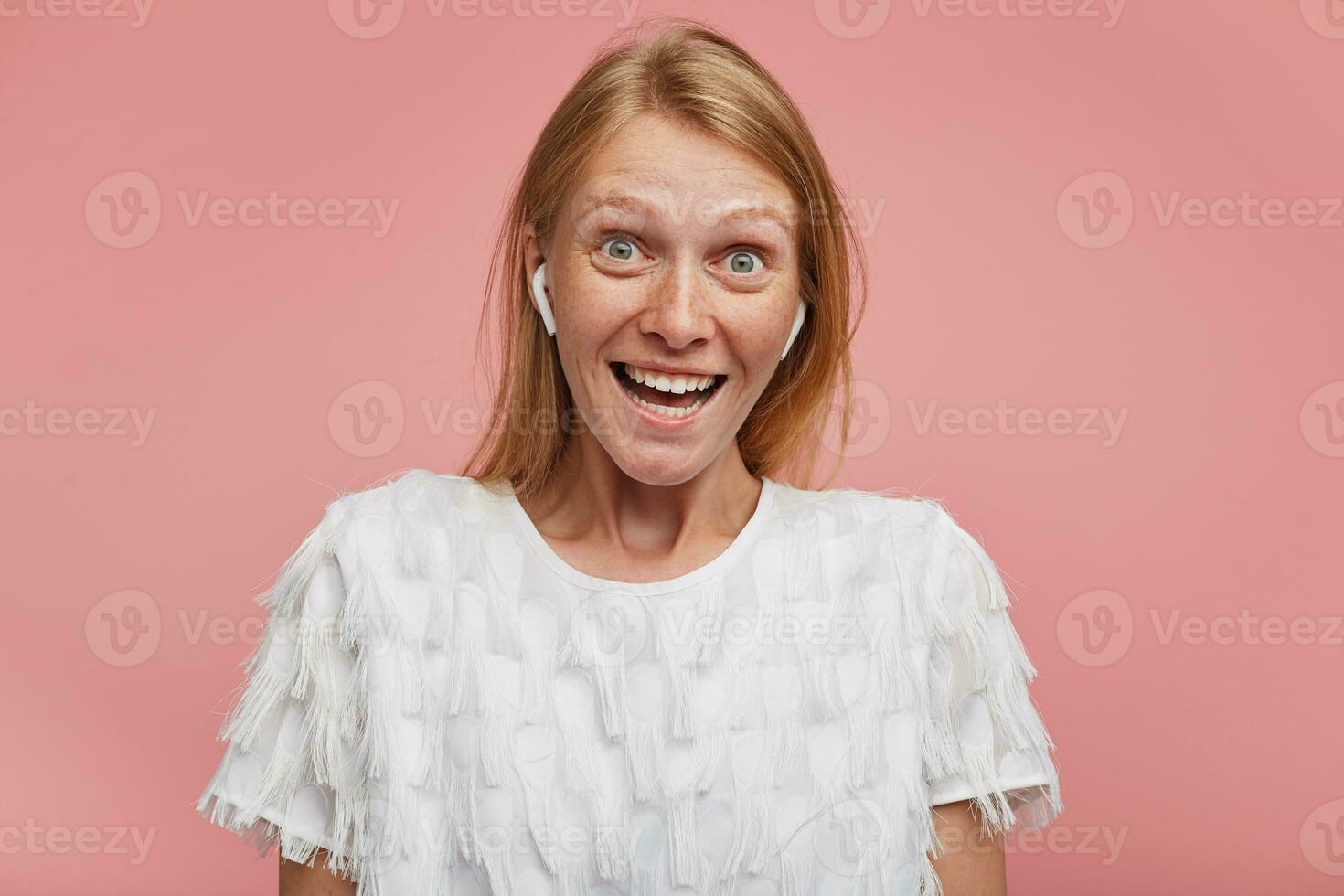 Overjoyed young pretty female with foxy hair raising surprisedly eyebrows while looking at camera with wide eyes and mouth opened, standing against pink background photo
