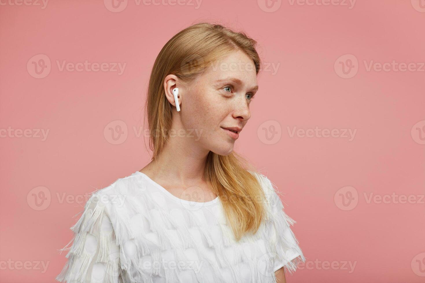 Studio photo of charming young positive redhead lady dressed in elegant clothes while posing over pink background with earphones, looking ahead and smiling gently