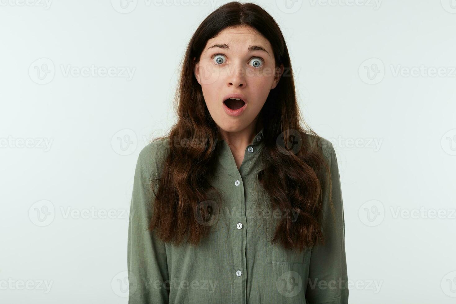 Shocked young long haired brunette female rounding amazedly her green eyes while looking surprisedly at camera, standing over white background with hands down photo