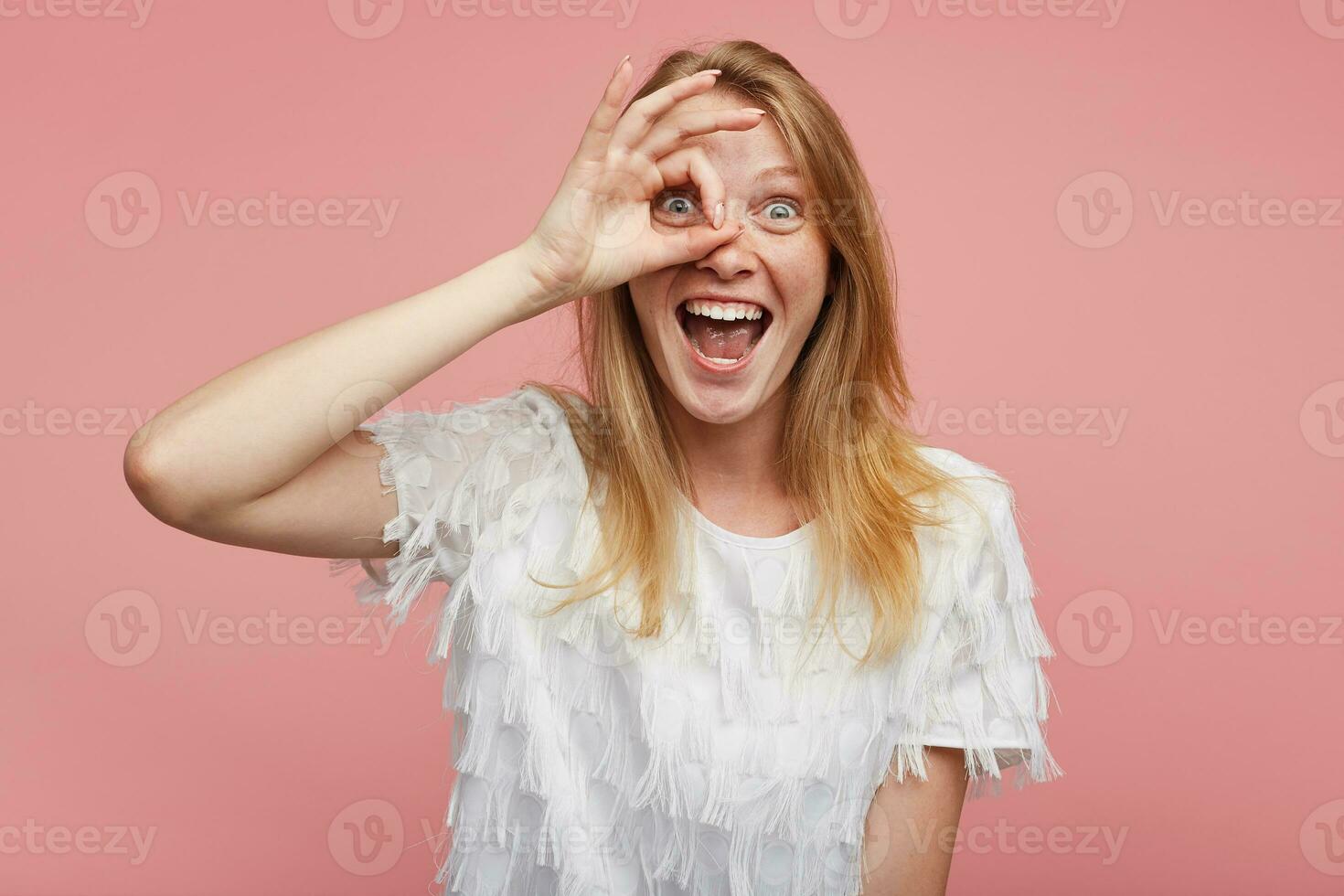 Joyful young beautiful woman with foxy hair raising ok gesture to her face and looking happily at camera and smiling happily with wide mouth opened, isolated over pink background photo