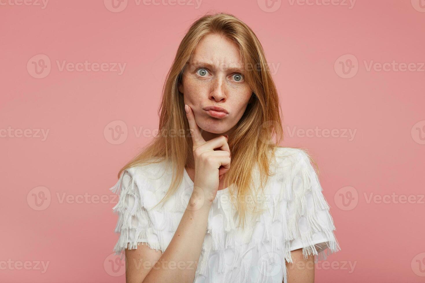 Amazed young pretty female with foxy hair rounding her green-grey eyes while looking at camera with pouted lips, holding index finger on her cheek while posing over pink background photo