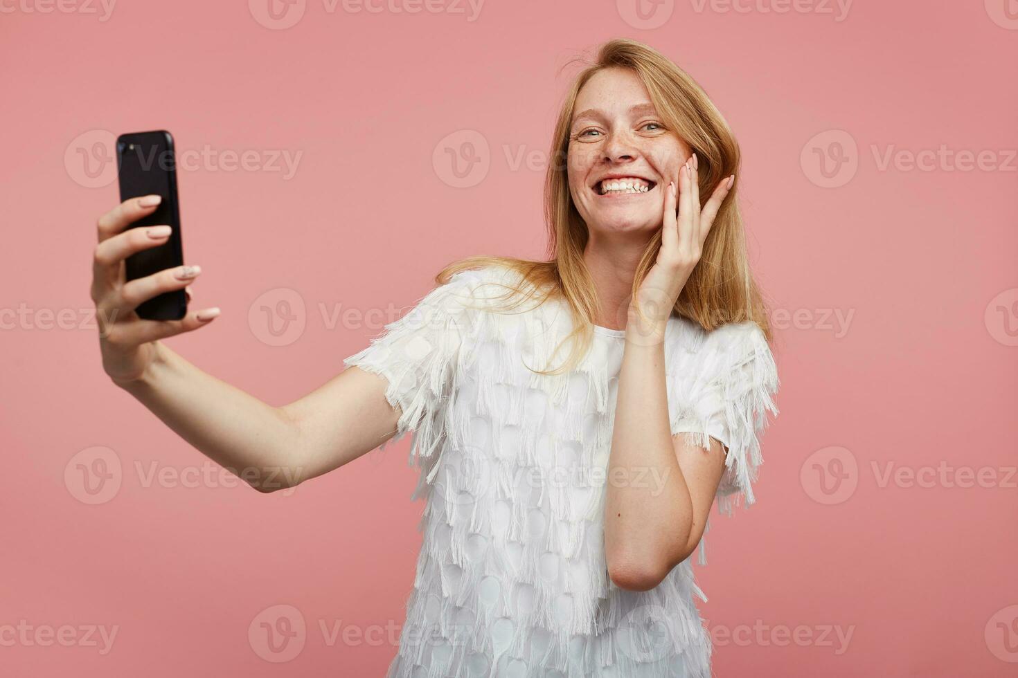Indoor shot of joyful young lovely female with foxy hair showing her white perfect teeth while smiling widely, making selfie with her smartphone while standing over pink background photo