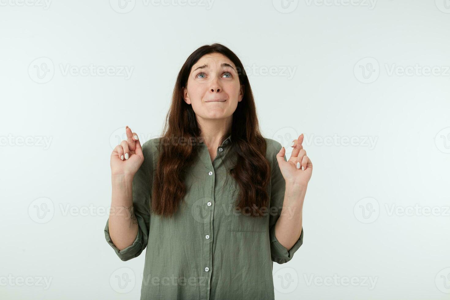 Hoping young attractive brown haired female with natural makeup keeping her fingers closed while looking worringly upwards, standing over white background photo