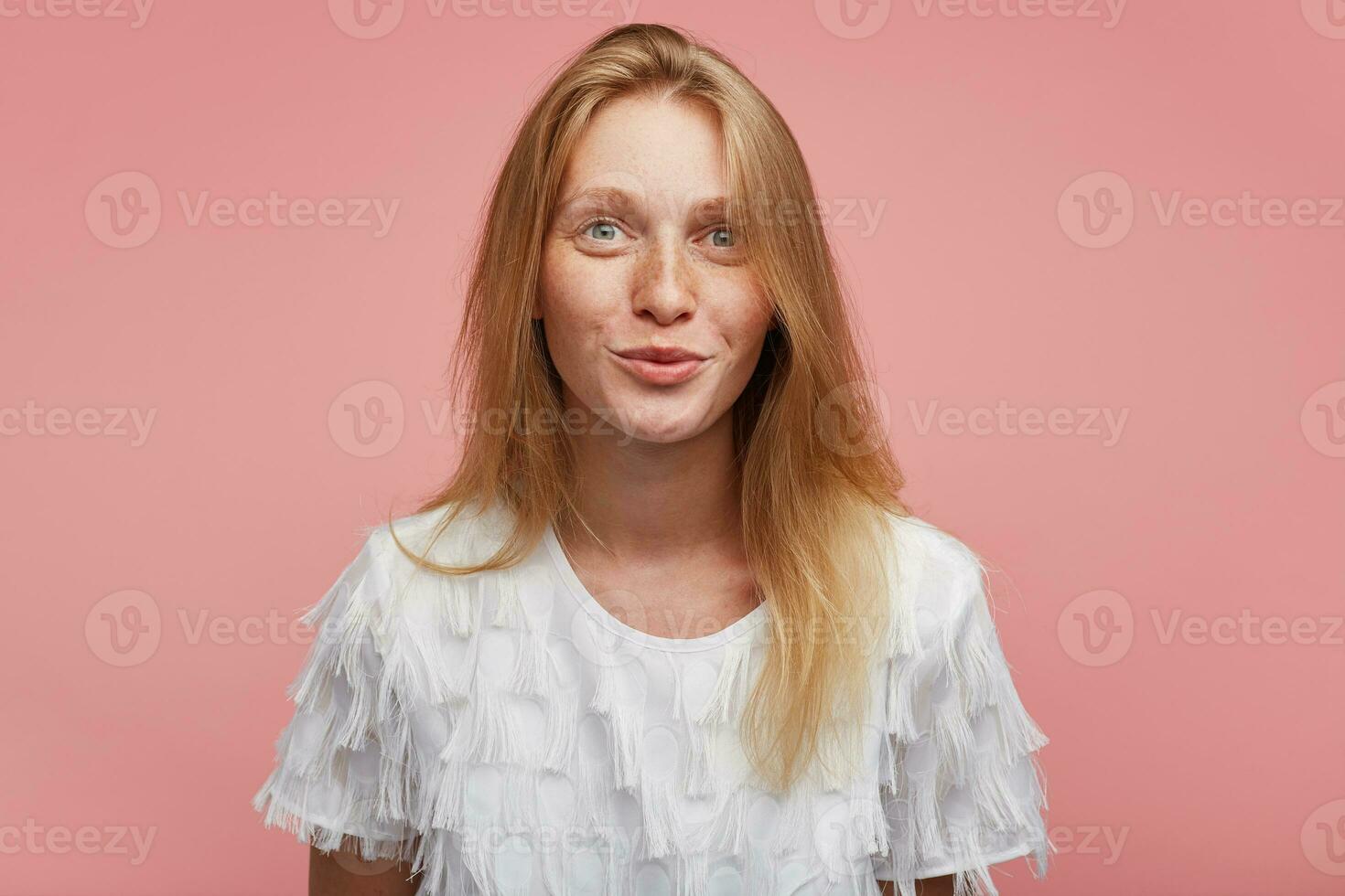 Portrait of attractive young redhead female with natural makeup looking positively at camera and smiling gently, standing over pink background with hands down photo