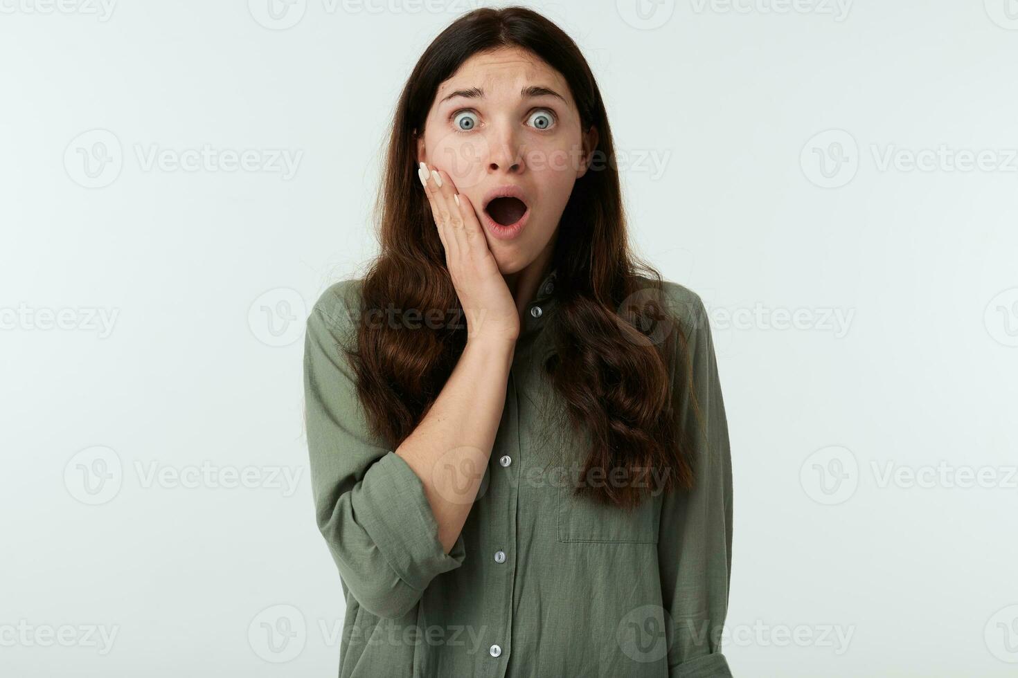 Astonished young pretty brown haired woman with natural makeup holding raised palm on her cheek while looking amazedly at camera with opened mouth, isolated over white background photo