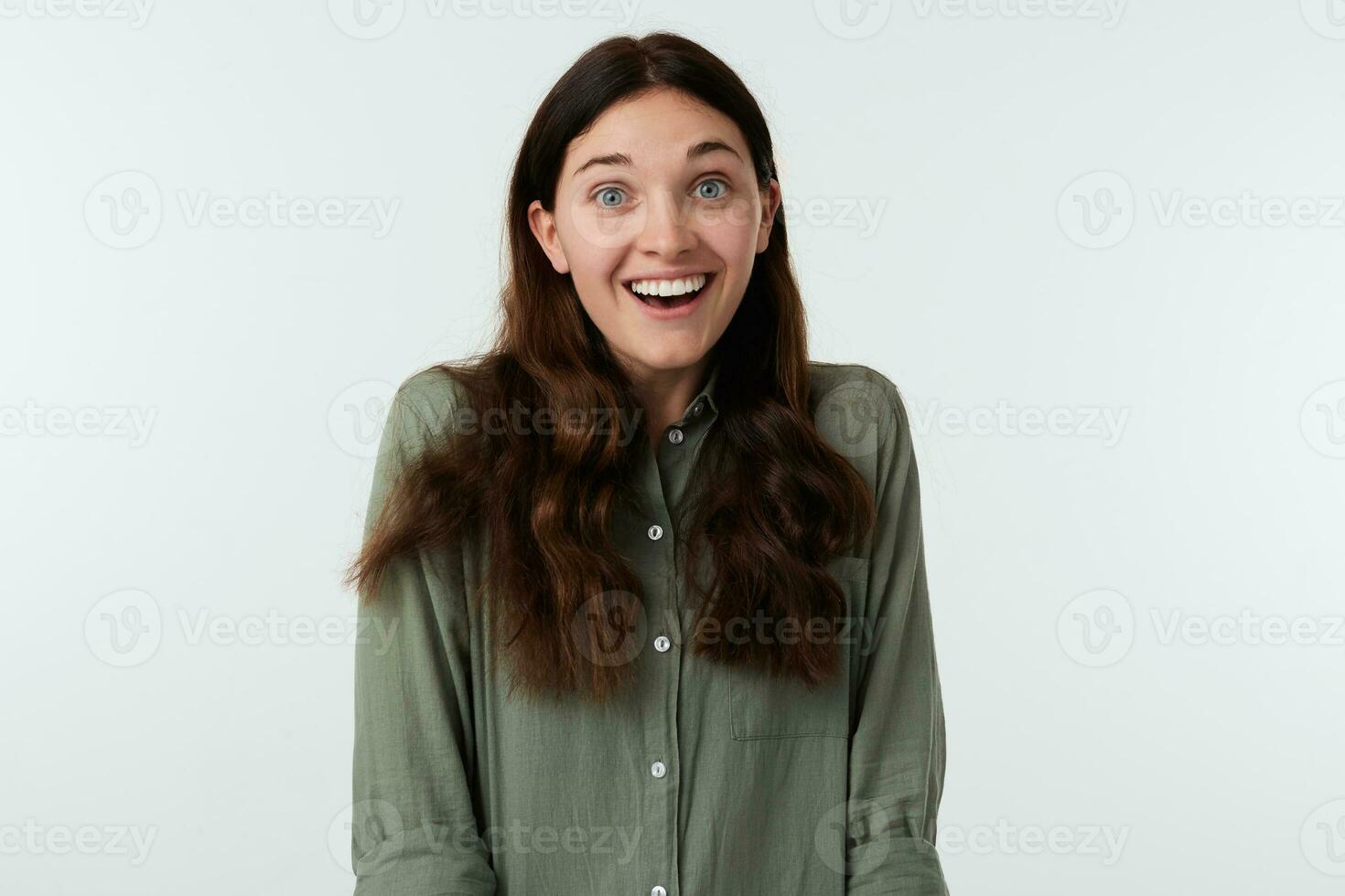 Overjoyed young lovely brown haired woman with natural makeup raising her eyebrows while looking cheerfully at camera with broad smile, isolated over white background photo