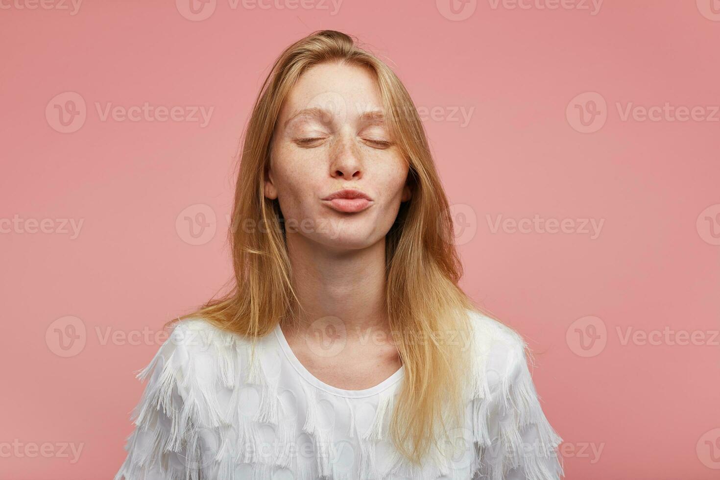 Pleasant looking positive lovely young lady with foxy hair folding lips in air kiss and keeping eyes closed while standing over pink background in elegant clothes photo